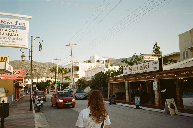 a woman is walking down the street in front of shops and cars parked on the side of the road