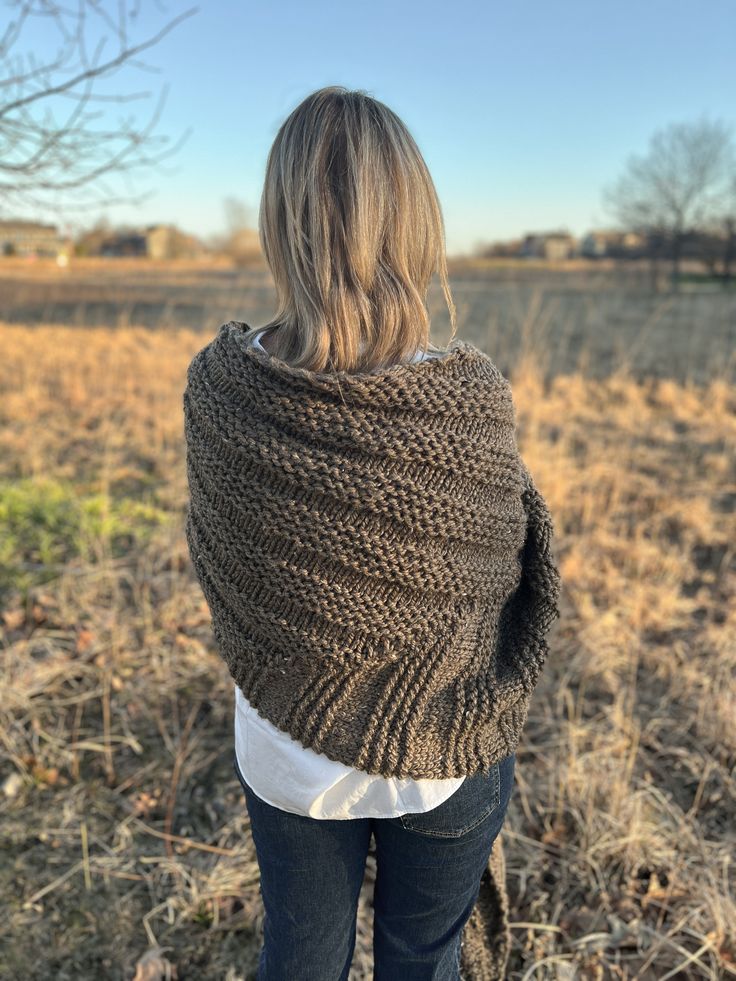 a woman standing in a field wearing a brown knitted shawl