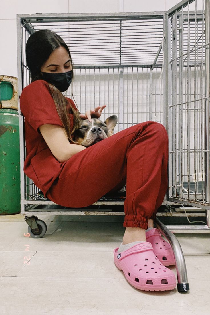 a woman sitting on the ground with her dog in a cage behind her, wearing a mask