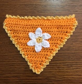 an orange crocheted triangle with a white flower on the front and bottom, sitting on a wooden surface