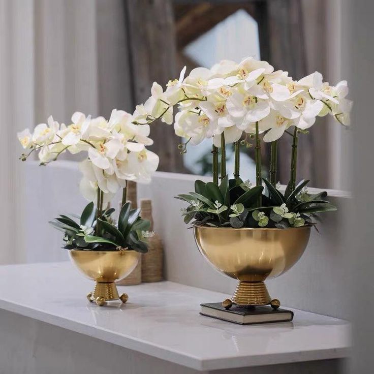 two gold vases with white flowers in them on a marble countertop next to a mirror