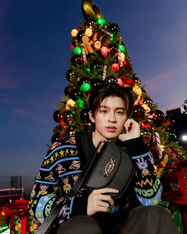 a young man sitting in front of a christmas tree holding a black bag and looking at the camera