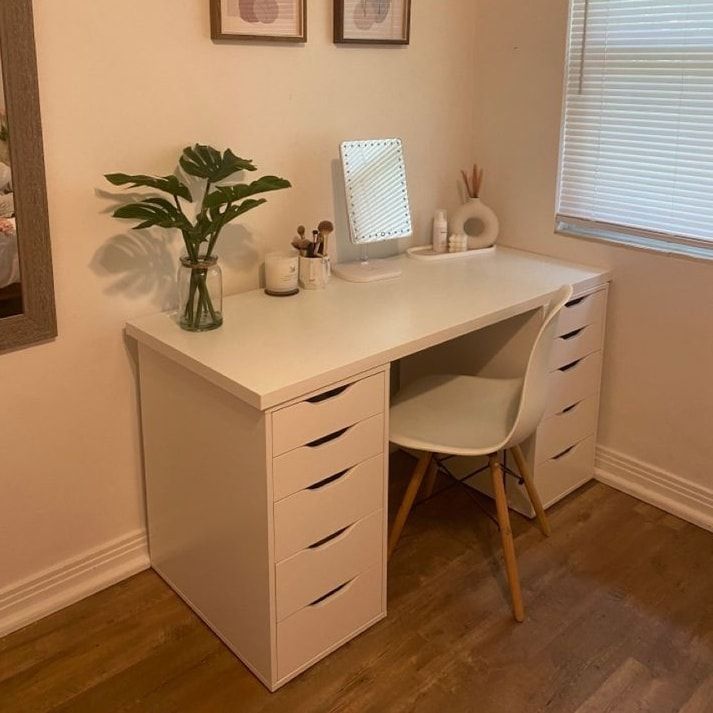 a white desk with two drawers and a plant on it in front of a window