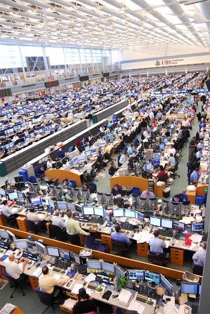 an office filled with lots of computers and people working at desks in front of large windows