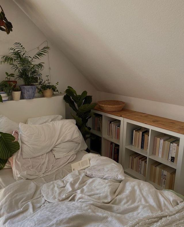 an unmade bed with white sheets and bookshelves in a room filled with potted plants