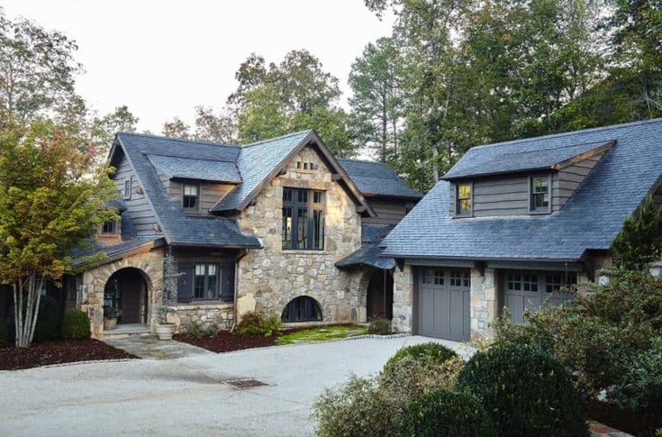 a large stone house with two garages and trees in the front yard, surrounded by shrubs