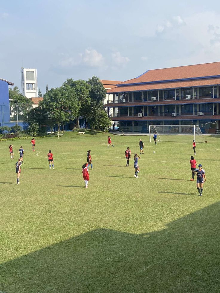 children are playing soccer on the grass in front of a school with buildings and trees