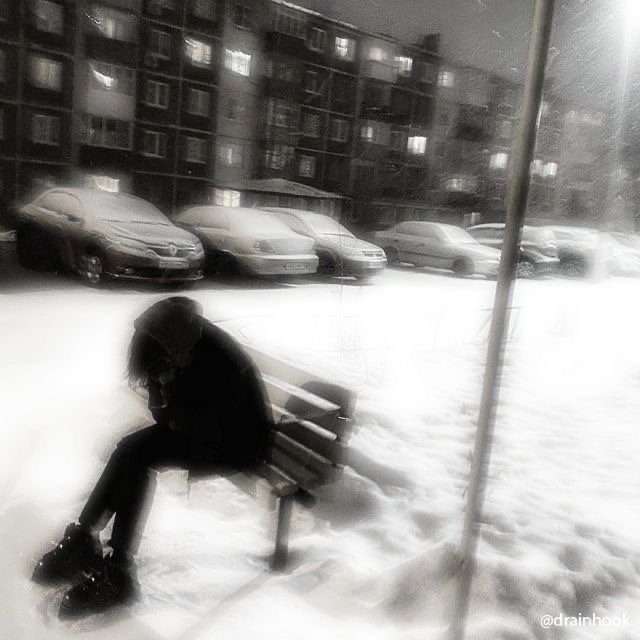 a person sitting on a bench in the snow next to some parked cars and buildings