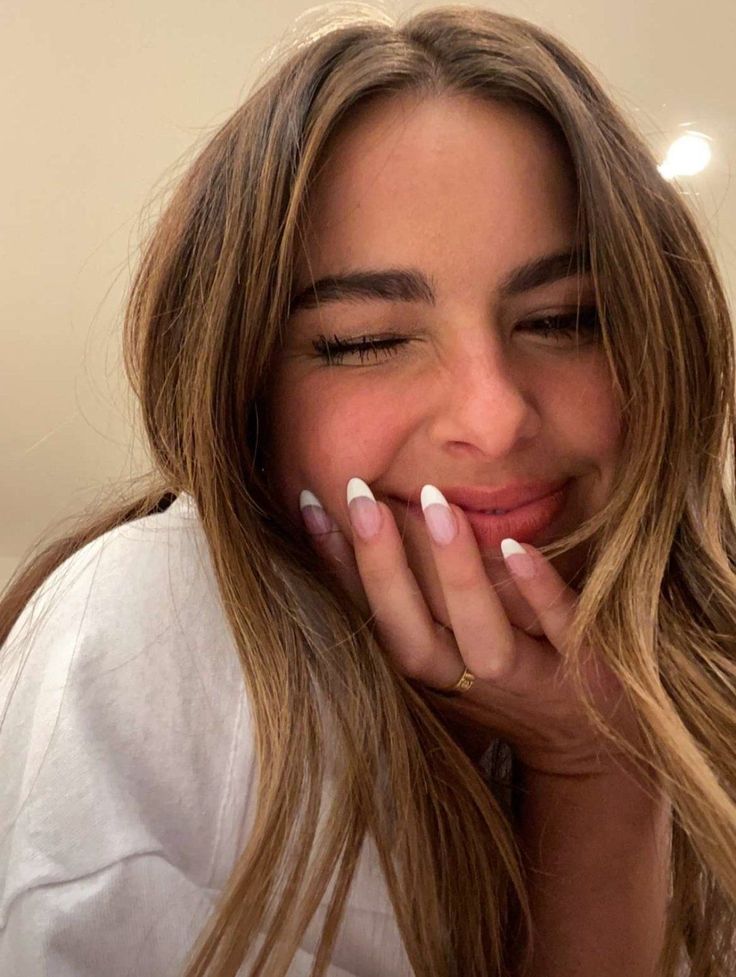 a woman with long blonde hair and white nails smiles while holding her hand to her face