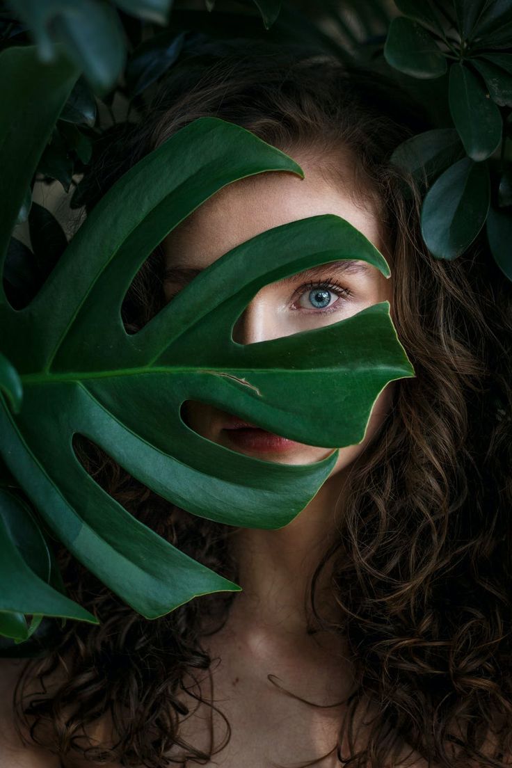 a woman with long curly hair and blue eyes is surrounded by green leaves in front of her face