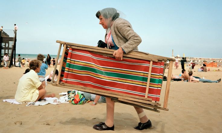 an old woman carrying a large blanket on the beach with people sitting around and standing in the sand