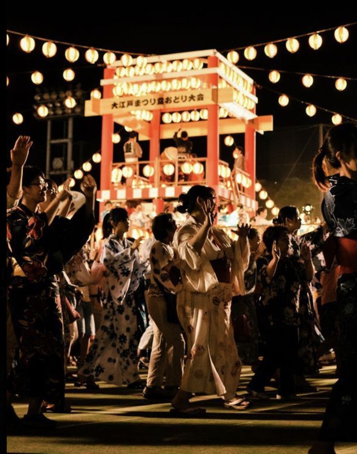 a group of people dancing in front of a building at night with lights strung above