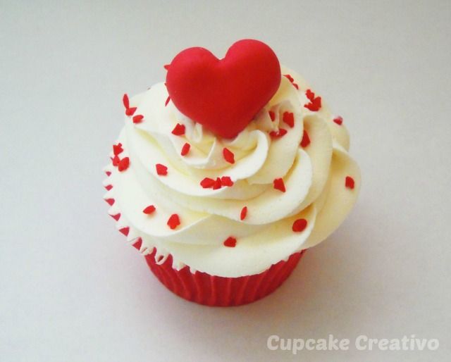 a cupcake with white frosting and red sprinkles has a heart on top