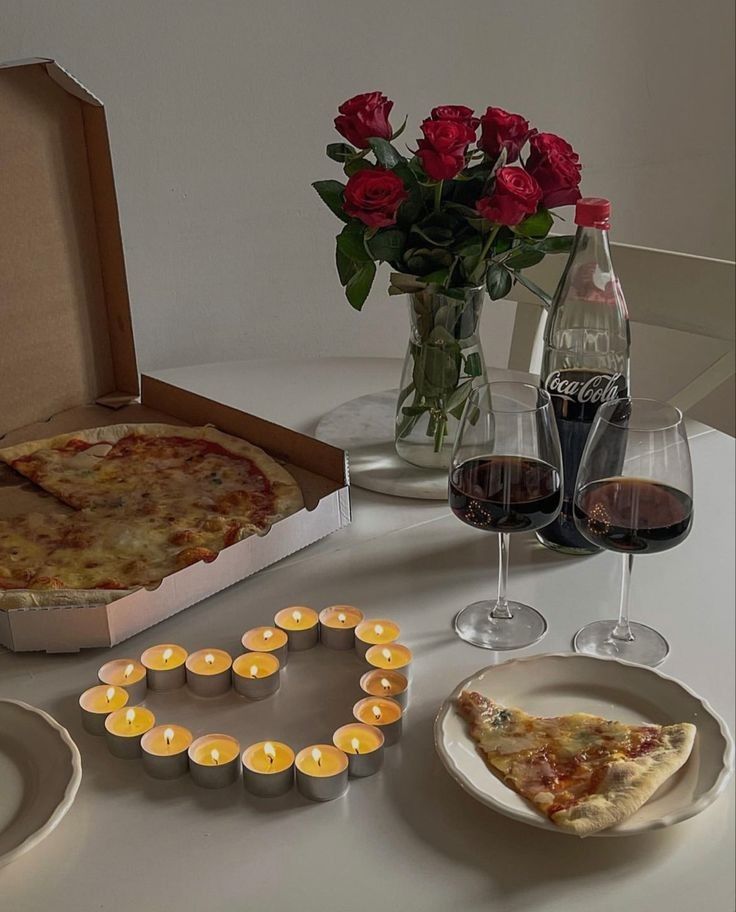 a table topped with two pizzas and glasses of wine next to a vase filled with roses