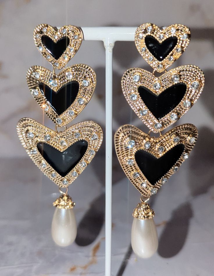 These stunning heart-shaped earrings feature three delicate hearts, gracefully dangling from a captivating gold outer interior with rhinestones for that extra sparkle. Adorn yourself with timeless beauty and express your love with every sway. Gold Heart Earrings With Rhinestones For Valentine's Day, Elegant Black Earrings With Heart Charm, Gold Rhinestone Heart Earrings, Gold Heart-shaped Earrings With Rhinestones, Elegant Black Metal Heart Earrings, Elegant Double Heart Earrings For Party, Glamorous Gold Heart Earrings, Elegant Double Heart Metal Earrings, Elegant Metal Earrings With Heart Charm