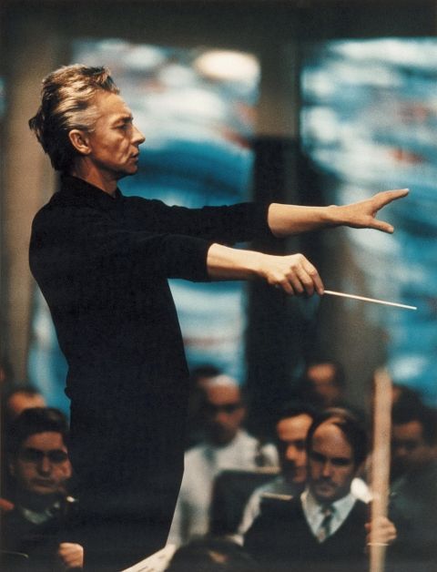 a man in black shirt and white pants holding a conductor's baton while people watch