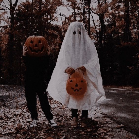 two people dressed up as ghost and pumpkins walking down a road with leaves on the ground