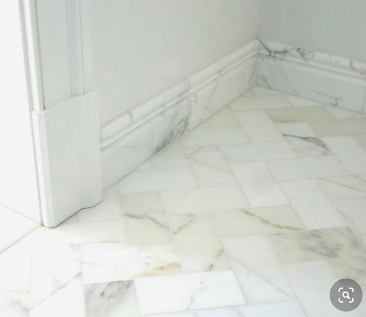 the corner of a bathroom with white marble flooring and wall tiles on the walls