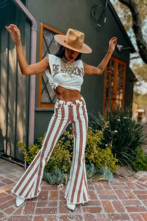 A person wearing a tied graphic crop top, the Dallas Striped Bellbottom Jeans in Rust by WISTERIA LANE, a wide-brimmed hat, and white shoes poses outdoors on a brick path with one arm raised and the other bent. A green building and plants are in the background. Nashville Style Outfits, Country Concert Outfits, Wisteria Lane, Cowgirl Style Outfits, Look Festival, Fest Outfits, Nashville Style, Western Wear Outfits, Cute Country Outfits