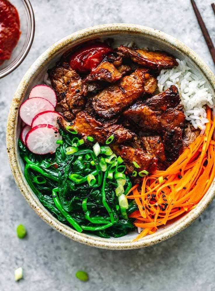 a bowl filled with meat and vegetables next to chopsticks on a table top