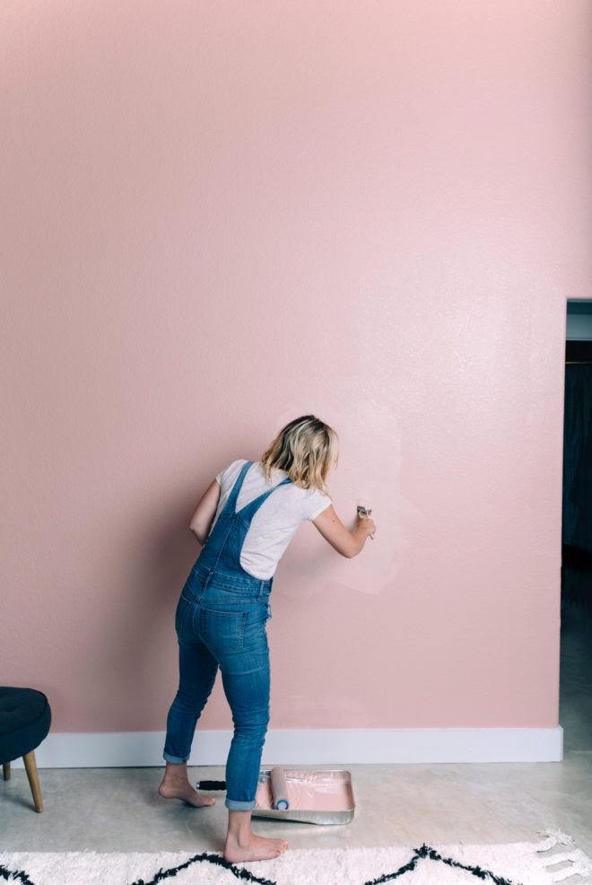 a woman in overalls painting a pink wall with a paint roller on the floor