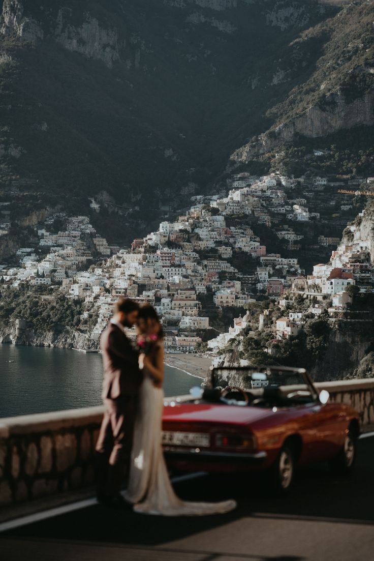 a bride and groom standing next to a red car in front of a mountain town