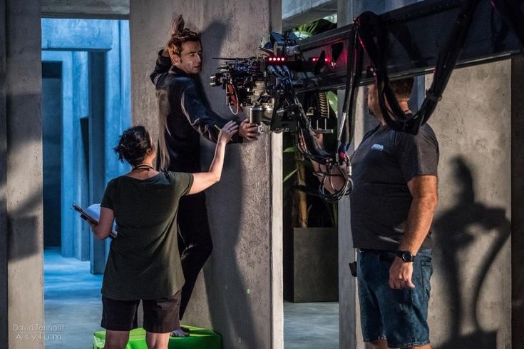 three people standing in front of a camera and another person on a stool looking at something