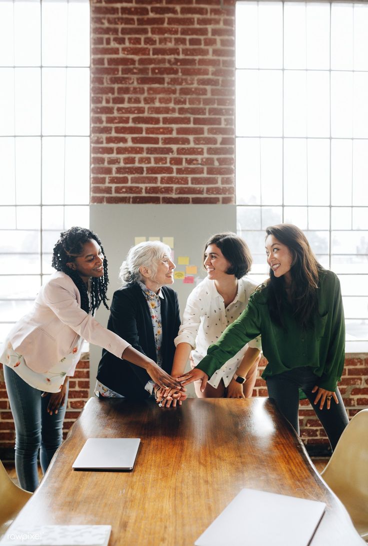 four women shaking hands over a wooden table in an office setting with brick walls and windows