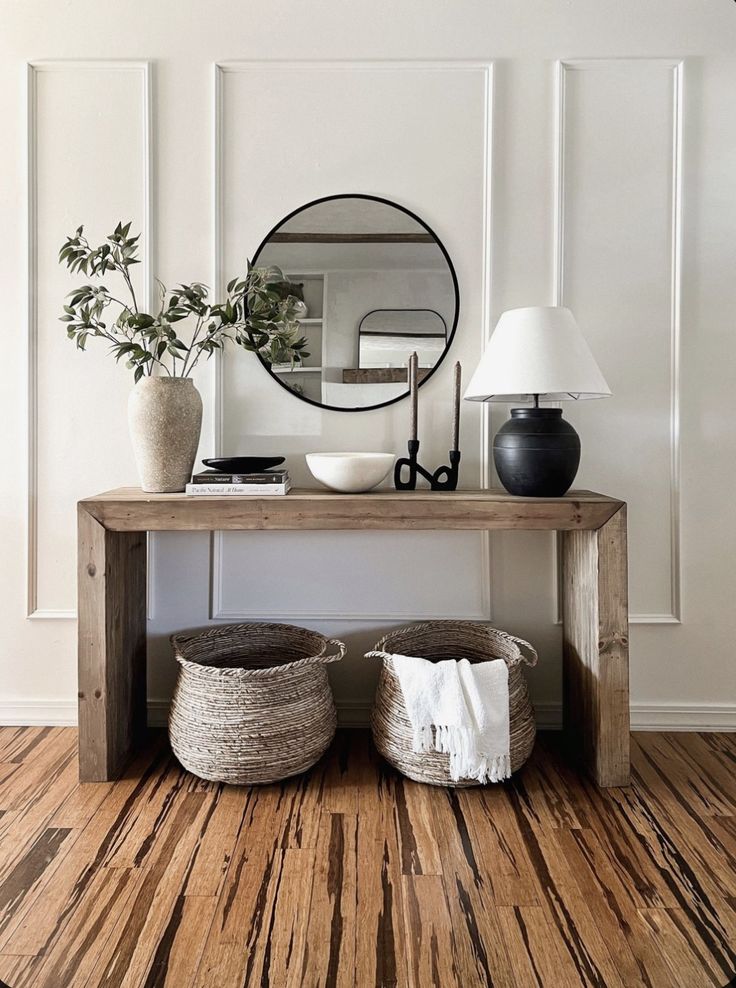 a wooden table topped with baskets next to a mirror and vase on top of a hard wood floor