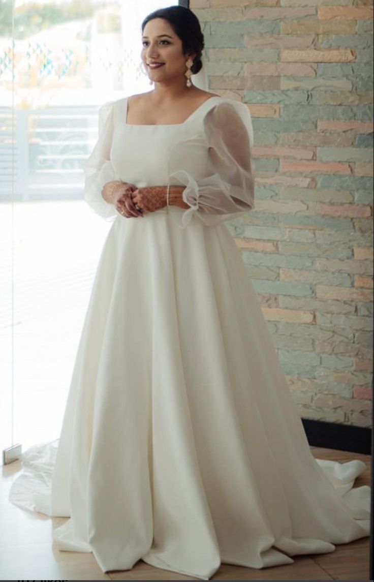 a woman standing in front of a brick wall wearing a white dress and long sleeves