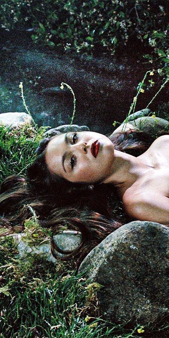 a naked woman laying in the grass next to some rocks and water with her eyes closed