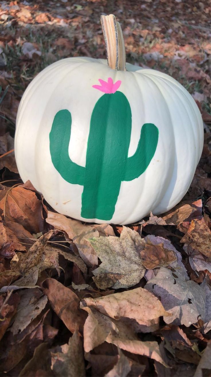 a white pumpkin with a green cactus painted on it sitting in the middle of leaves