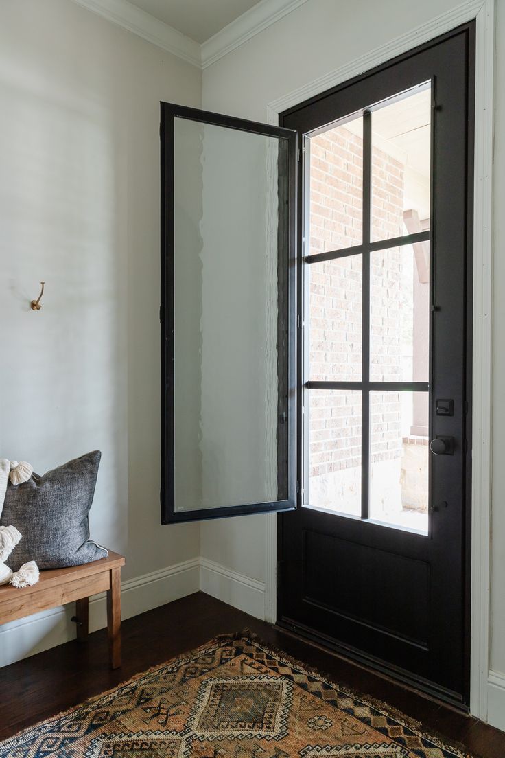 a black door is open in front of a white wall and wooden bench with pillows on it