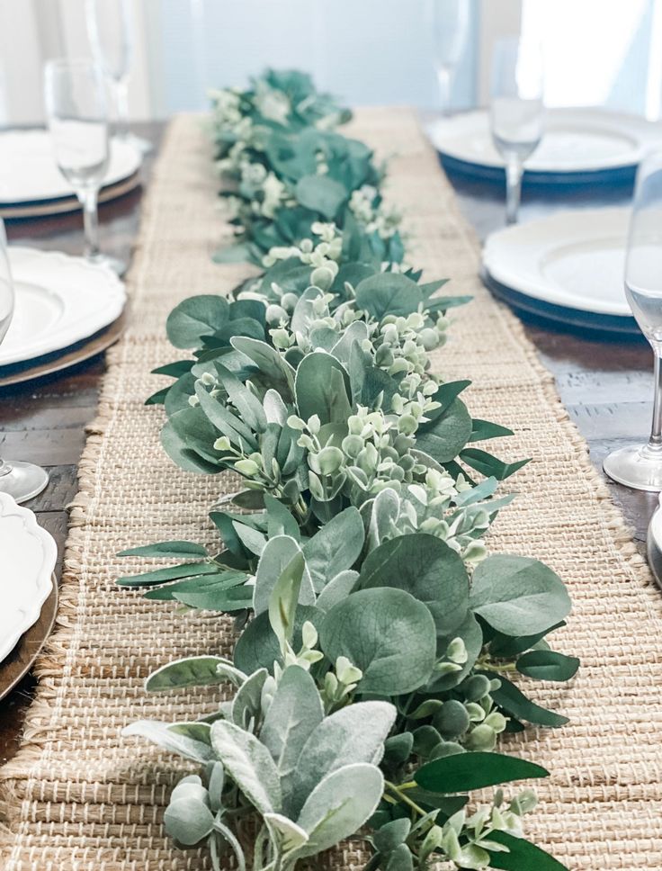 the table is set with white plates and place settings, along with greenery on each side