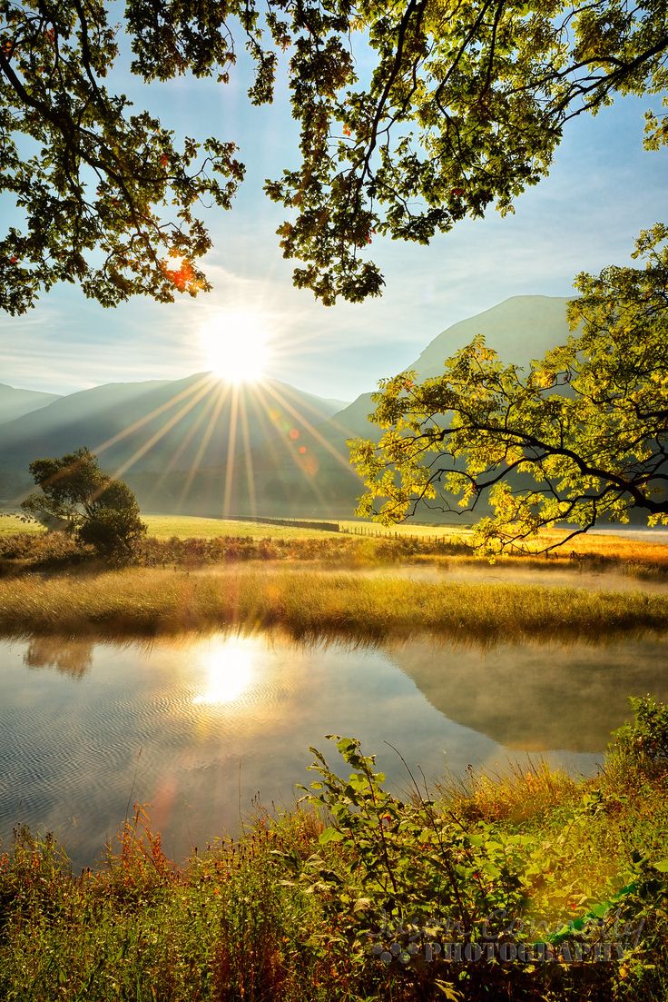 the sun shines brightly over a small lake surrounded by trees and grass, with mountains in the background