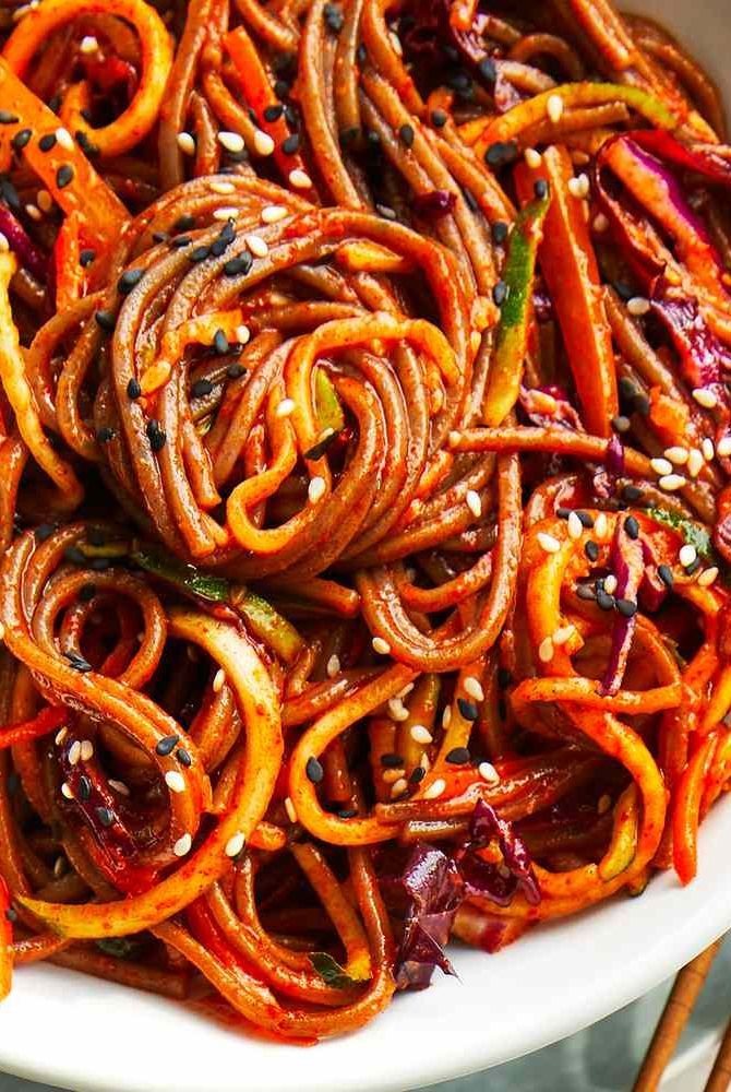 a white bowl filled with stir fry noodles covered in sesame seeds and carrots next to chopsticks