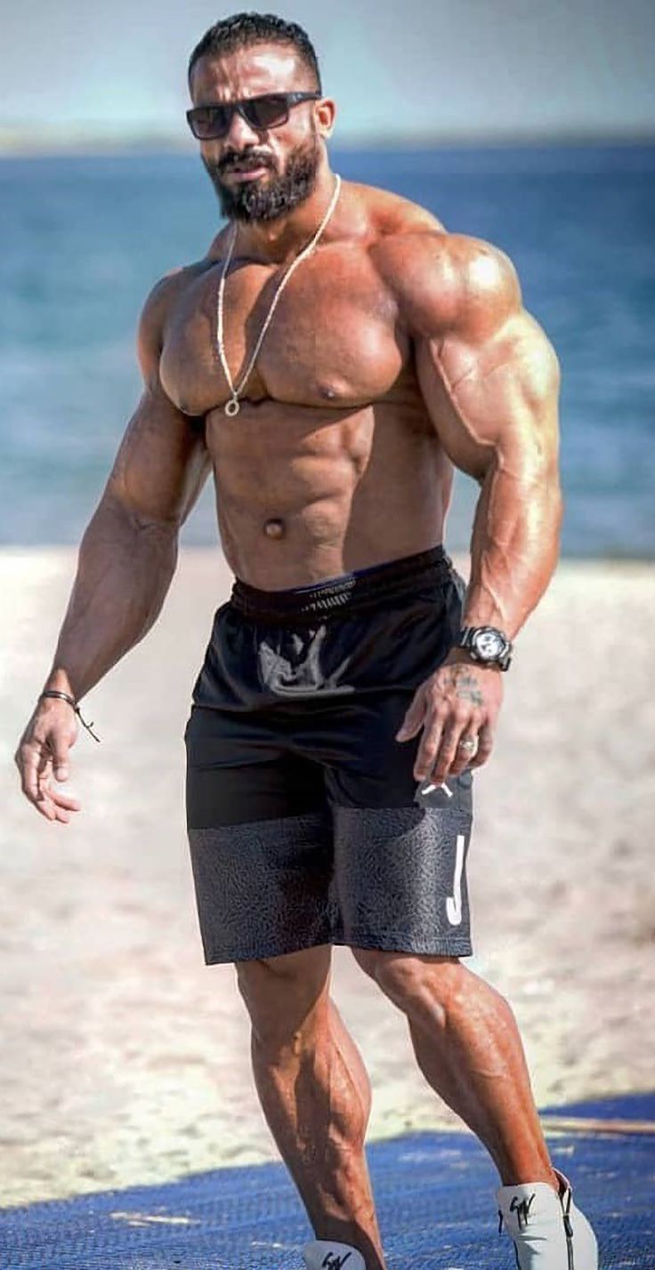 a shirtless man standing on the beach with his hands in his pockets and looking at the camera