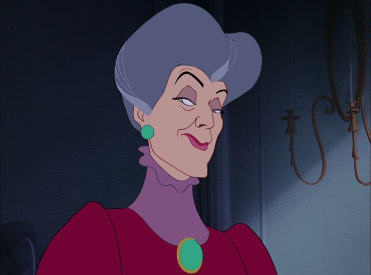 an older woman with grey hair and green earrings looks at the camera while standing in front of a chandelier