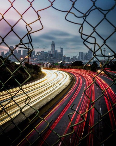 a city skyline is seen through a chain link fence with traffic lights streaking by