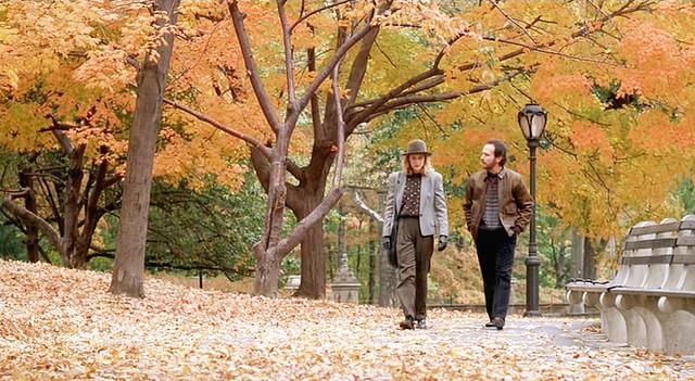 two people are walking in the park with leaves on the ground and one is wearing a hat