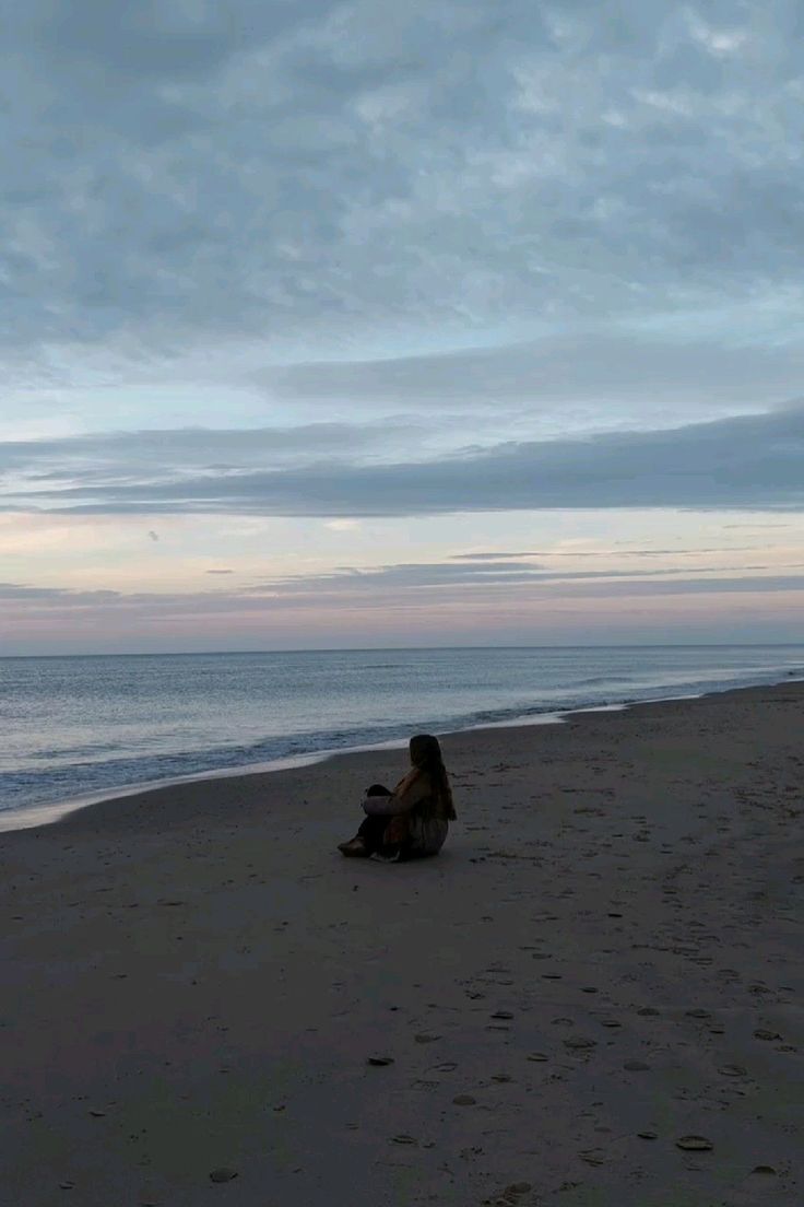 a woman sitting on top of a sandy beach next to the ocean under a cloudy sky