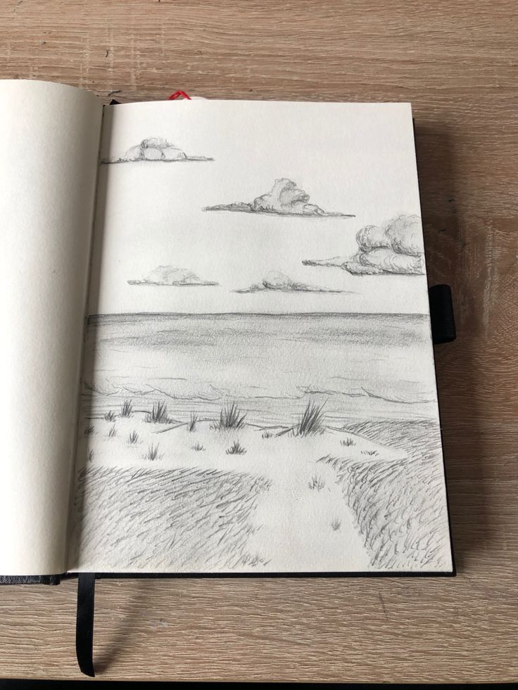 an open notebook with a drawing of a beach and clouds in the sky, on top of a wooden table