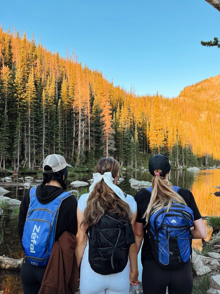 three people with back packs are walking towards a lake in the mountains near some trees