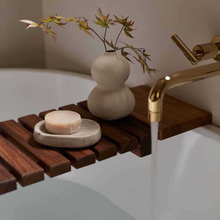 a white vase with flowers and soap on a wooden shelf next to a bathtub