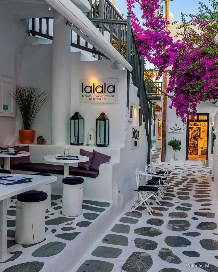 the outside of a restaurant with tables and chairs under purple bougaia trees in front of it