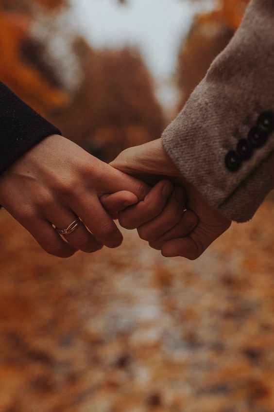 two people holding hands while walking down a path in an autumn forest with leaves on the ground