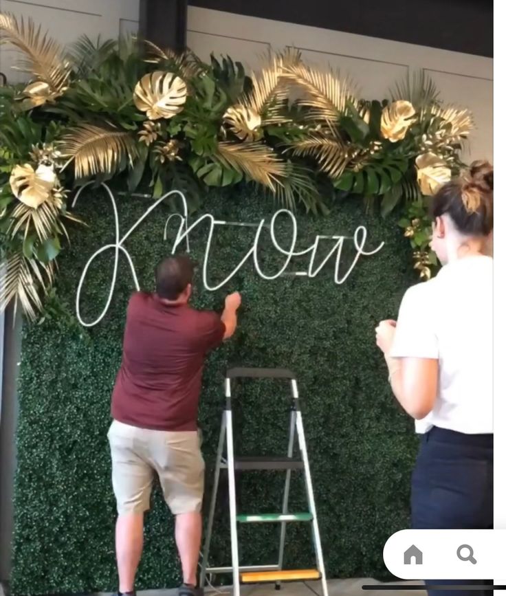 two people writing on a wall with flowers and greenery behind them, while another person stands on a ladder to paint the word