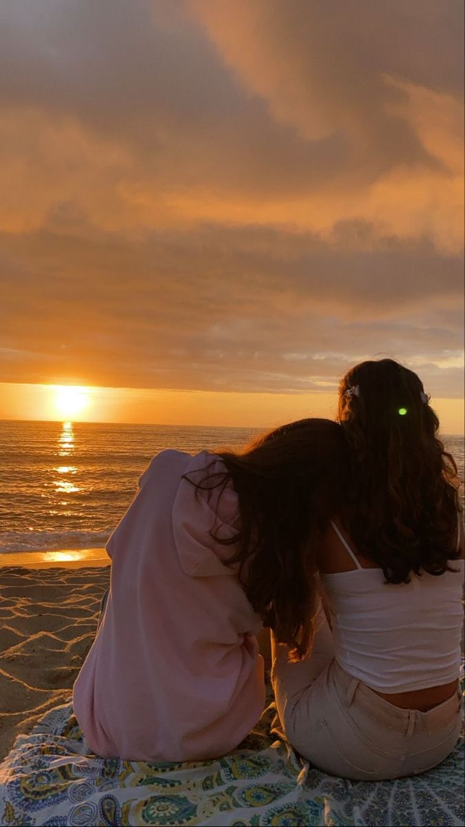 two women sitting on the beach watching the sun go down over the ocean at sunset