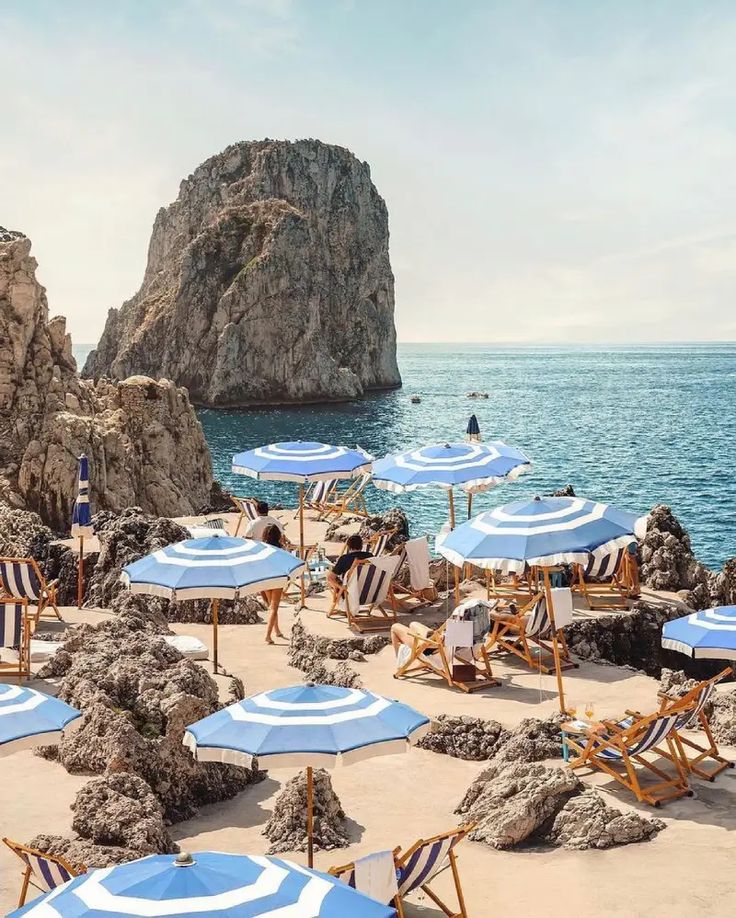 many beach chairs and umbrellas on the sand by the water with rocks in the background