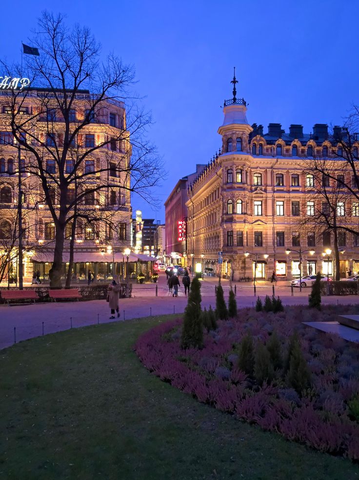 people are walking around in front of some buildings and trees at night with the lights on